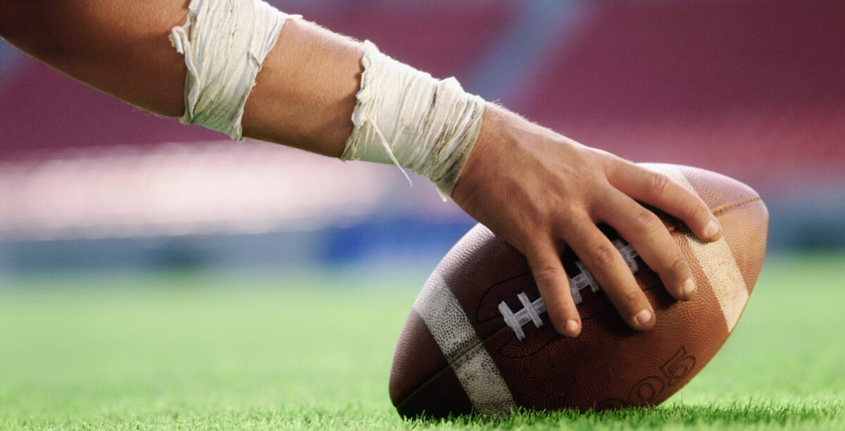 A hand holding a football down on the turf of the football field
