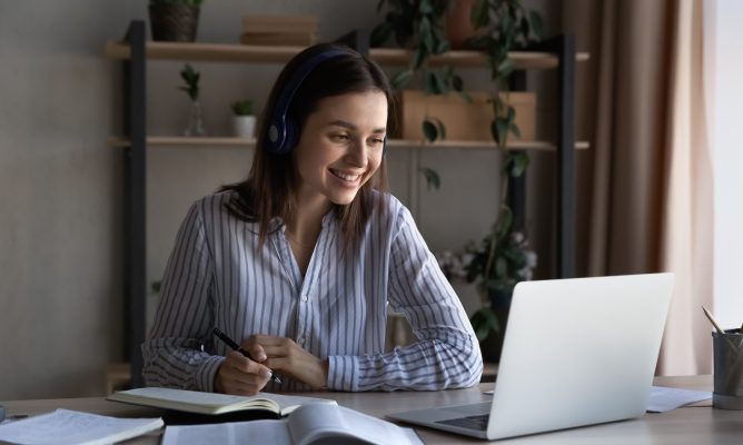 Happy woman wearing headphones while watching a webinar and taking notes