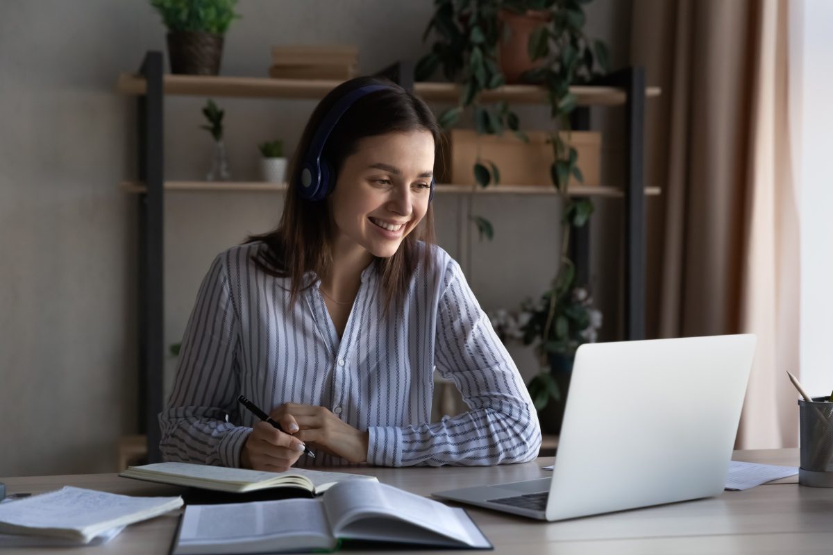 Happy woman wearing headphones while watching a webinar and taking notes