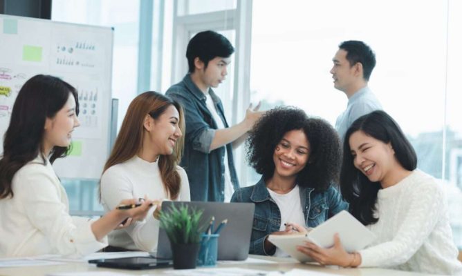 Four women working and talking around a computer with two men talking in the background
