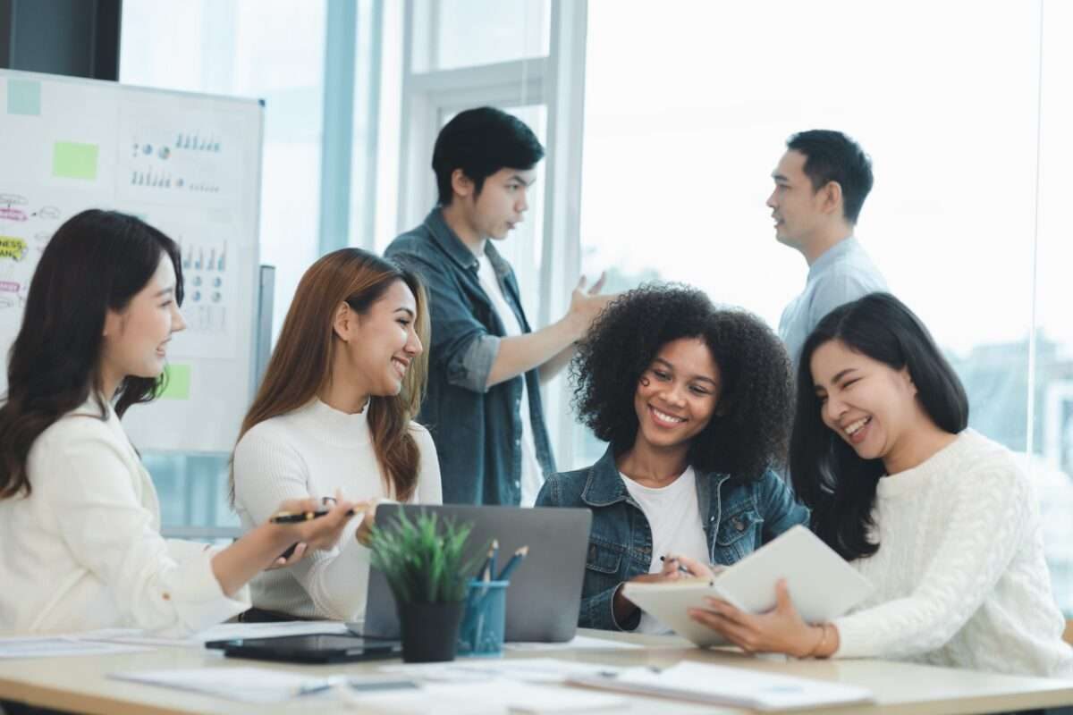 Four women working and talking around a computer with two men talking in the background