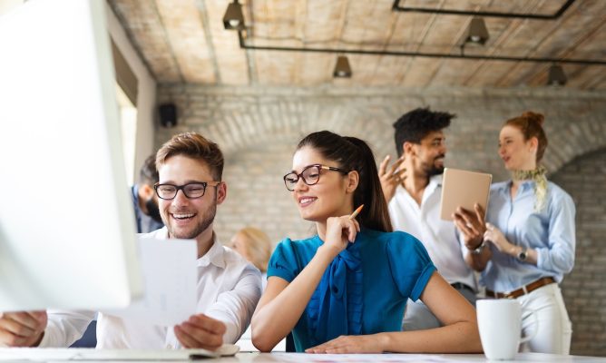 Coworkers working on the computer together with a male and female coworker talking in the background
