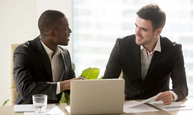 Two businessmen sitting at a desk while talking to each other