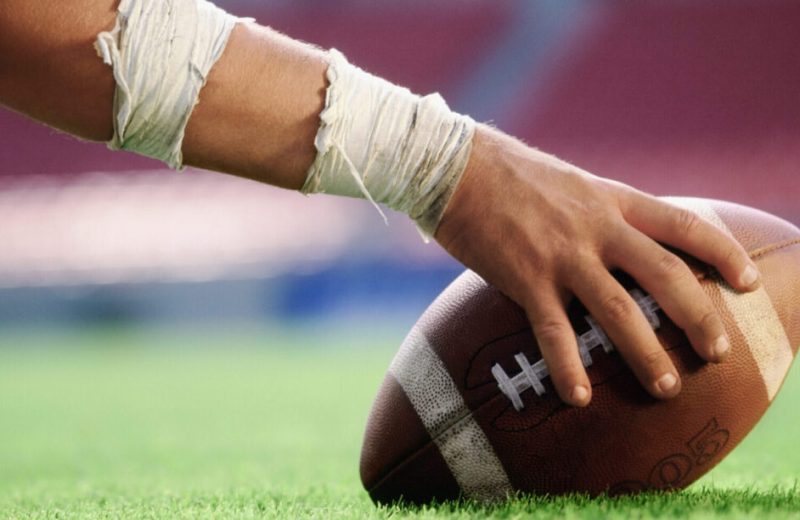A hand holding a football down on the turf of the football field