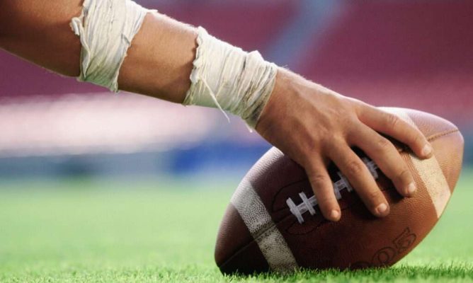 A hand holding a football down on the turf of the football field