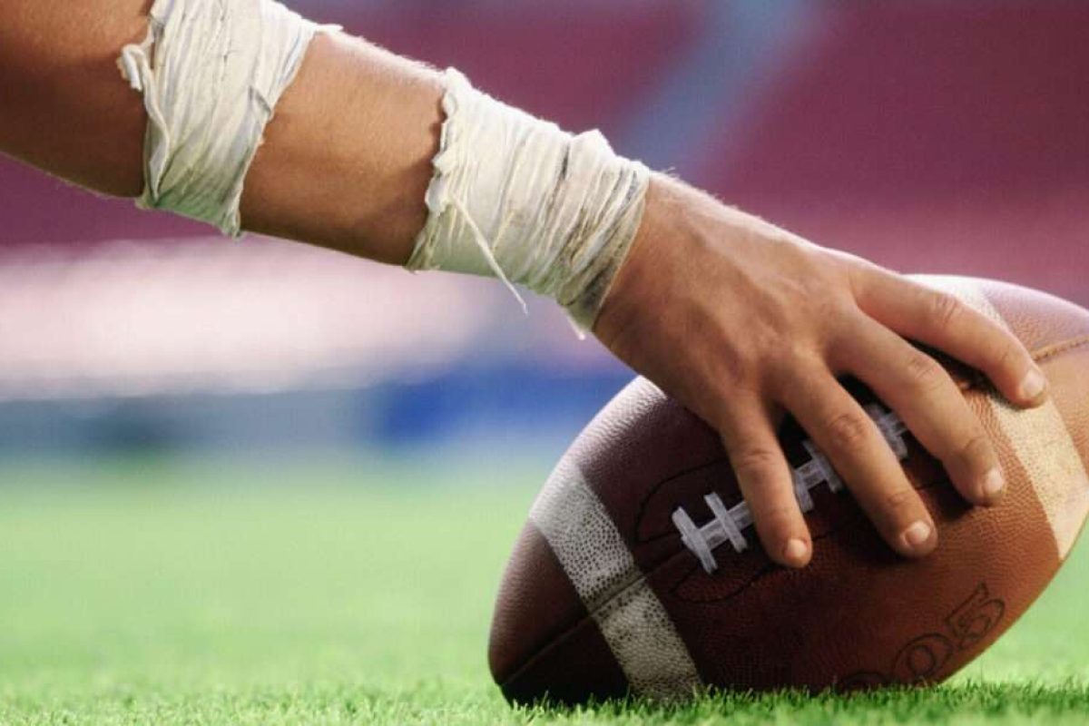 A hand holding a football down on the turf of the football field